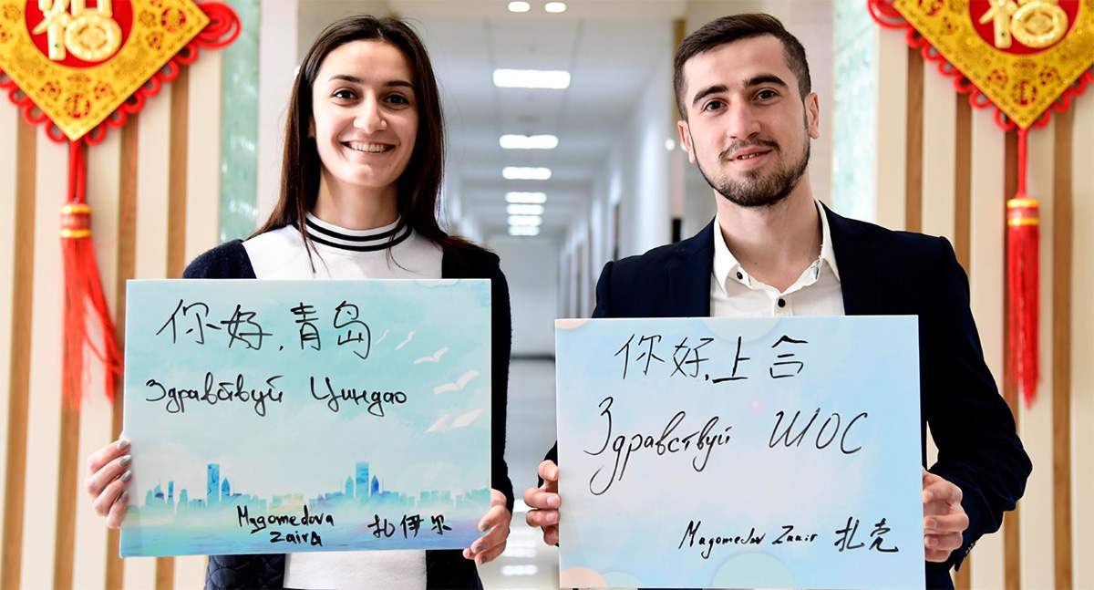 Хятадын Газрын тосны их сургуульд сурдаг Оросын оюутан Закир (баруун гар талаас), түүний эгч Зайра нар “Сайн байна уу, Циндао”, “Сайн байна уу, ШХАБ” гэж хятад болон оросоор бичин барьжээ. 2018 оны тавдугаар сарын 3-нд Циндао хотод. (Гэрэл зургийг Синьхуа/Гуо СюйЛэй)